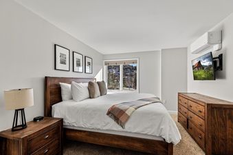 A serene bedroom at Bear Claw 2 Valley View 207 featuring a wooden king-sized bed with white linens and a plaid throw, a matching nightstand with a lamp, a wooden dresser, a wall-mounted TV, an air conditioning unit, and a window with a valley view.