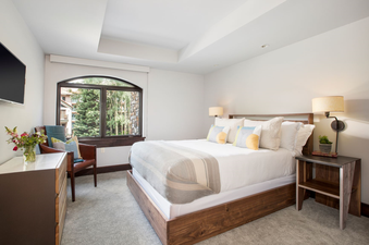 The bedroom in Lumiere Three Bedroom Unit 2 Bath features a large, comfortable bed with white linens and a grey throw, bedside tables with lamps, and a large window with a view of the trees outside.