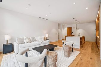A modern open-plan living room and kitchen in Founders Place 2401, featuring a white sofa, cream armchair, dark coffee table, and a kitchen island with pendant lights.