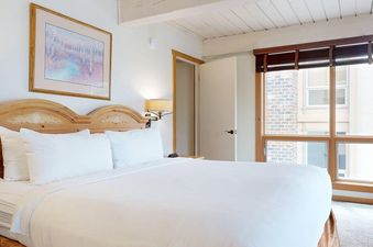 A king-sized bed with white linens and a wooden headboard is featured in the bedroom of Top of the Village Slope 310, with a window on the right side offering a view of the surrounding buildings.