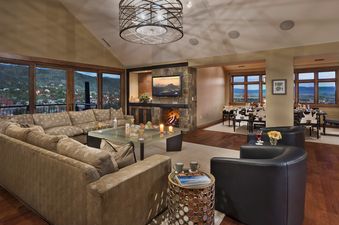A luxurious and spacious living area, part of Flat Tops Peak Penthouse One Steamboat Place 701, features a large U-shaped sofa surrounding a glass coffee table, with a lit fireplace and flat-screen TV on a stone accent wall, and a dining area visible beyo