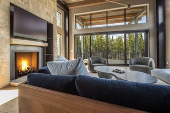 A cozy living room in Little Thunder Chalet features a fireplace with a roaring fire and a large television above it, comfortable sofas, and expansive windows offering a view of a lush forest.