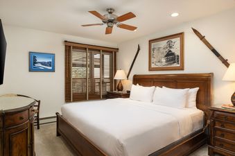 A cozy king bedroom at Montaneros 318, featuring a king-sized bed with white linens, wooden nightstands, elegant lamps, and rustic art, with a ceiling fan and large window with wooden blinds.