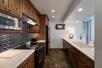The galley kitchen at Vails Mountain Haus Three Bedroom Valleyside features wooden cabinetry, white countertops, and black and white patterned backsplash.