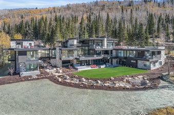 An aerial view of The Foundry, a sprawling modern mountain home with extensive glass and stone, surrounded by autumn trees and featuring a large green lawn.
