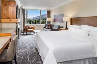 A luxurious and spacious hotel room features a large bed, a comfortable seating area, and a stunning view of snow-capped mountains and evergreen trees through a wide window.