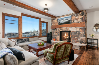 The living room at Grand Lodge Three Bedroom 301 has a fireplace, a large sectional sofa, and floor-to-ceiling windows offering stunning views of the mountains.