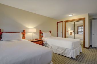 A cozy bedroom features two twin beds with white linens, a wooden nightstand with a lamp, and a closet with mirrored doors, all under warm lighting.