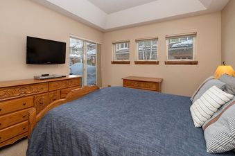 The bedroom at Telemark 514 features a spacious blue bed, a wooden dresser with a TV mounted above it, and large windows revealing a snowy outdoor scene.