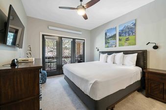 A master bedroom at The Charter A2505 featuring a king-sized bed, ceiling fan, wall-mounted TV, and glass French doors leading to a private balcony.
