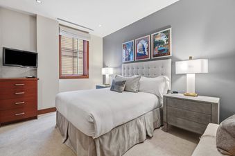 A cozy bedroom with a queen-sized bed, two bedside tables with lamps, a dresser with a TV, and a window with blinds.