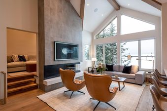 A luxurious living room in Northern Lights 32 featuring a large fireplace with a TV mounted above, two brown leather armchairs, a grey sofa, and expansive windows offering a snow-covered mountain view.