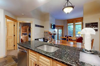 The kitchen at Taylors Crossing 109 features dark granite countertops, a stainless steel sink, and light wood cabinetry, with a cozy living area visible in the background.