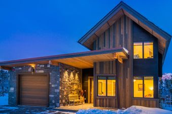 The Moonlight Basin Lodging 7 Rolling Ridge is a modern rustic home with warm lighting, set against a twilight sky and snowy landscape.