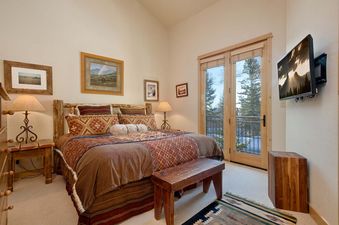 A rustic bedroom in Moose Creek 14 with a log bed, warm bedding, a wall-mounted TV, and glass doors opening to a snowy forest view.