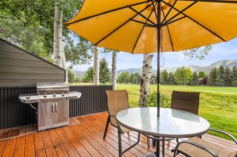 An outdoor deck with a BBQ grill, dining table with a yellow umbrella, and chairs, overlooking a golf course and mountains under a blue sky.