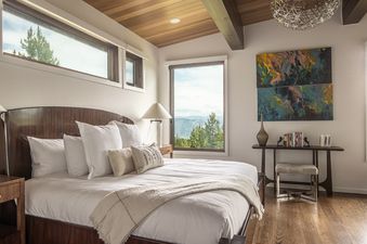 A luxurious bedroom at Yeti Ridge Retreat features a large bed with a wooden headboard, white linens, and pillows, with mountain views through wide windows, and abstract art above a console table.