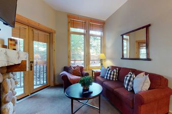 A cozy living room at Taylors Crossing 109 with a red sofa, armchair, and a stone fireplace, offering a view of the outdoors through large windows.