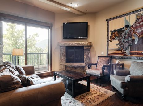 A cozy living room in 103 Juniper Landing features a large brown sofa, a stone fireplace, a wall-mounted TV, and a decorative tapestry on the wall.
