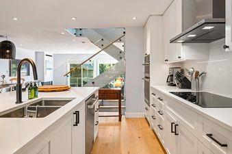 A modern kitchen with white cabinets, stainless steel appliances, and a large island with a sink, leading to a staircase with glass railings.