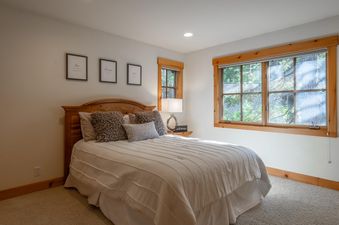 A serene bedroom at Northstar Sierra Gold Home featuring a comfortable bed, inspirational wall art, a bedside lamp, and large windows with natural wood frames offering views of lush greenery.