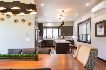 A modern kitchen and dining area in Sky High Colorado featuring dark wood cabinetry, a large island with gold fixtures, a dark wood dining table with leather chairs, and contemporary lighting.