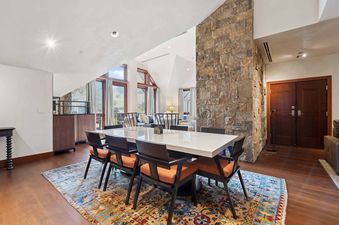 A spacious dining area with a modern white table, surrounded by stylish chairs with orange cushions, set on a colorful patterned rug.