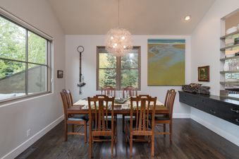 A modern dining area with a wooden table, chairs, a unique light fixture, and windows with green tree views.