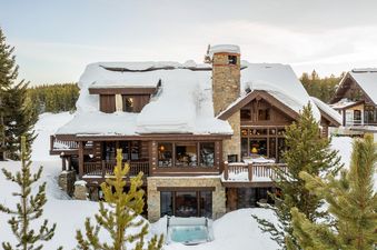 A large, multi-story mountain home, the Spanish Peaks Nestledown, is nestled among snow-covered evergreens under a clear winter sky, featuring a stone chimney and a hot tub sunken into the snow on the lower level.