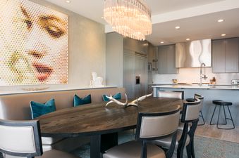 A modern dining room and kitchen area with an oval wooden dining table, light-colored dining chairs, a built-in banquette with teal pillows, and a large, colorful artwork on the wall, leading into a sleek gray kitchen with stainless steel appliances and a