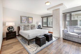 A modern bedroom with a king-sized bed, two nightstands with lamps, a blue and cream area rug, two ottomans, and a gray sofa.