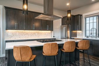 A modern kitchen with dark wood cabinets, a large island with a stovetop, and four leather bar stools.