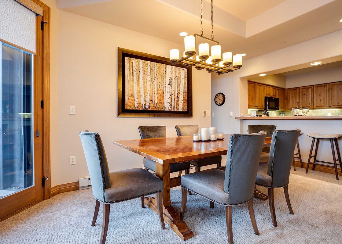 A sophisticated dining area featuring a wooden table with six upholstered chairs, illuminated by a rectangular chandelier, with a forest artwork on the wall and a partial view of the kitchen.