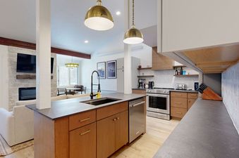 The Hollyhock 2023 kitchen features an island with a dark countertop and a stainless steel dishwasher, with a kitchen area on the right, and a living room and dining room in the background.