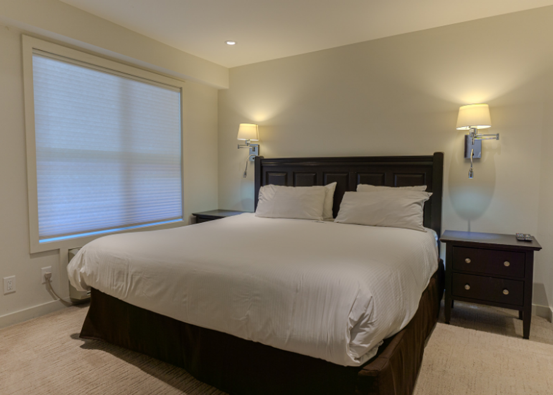 The bedroom at Aspens on Blackcomb Unit 451 features a comfortable bed, a window with blinds, and nightstands with lamps on either side.
