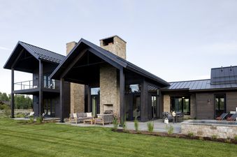 A modern luxury home with dark wood siding and stone accents features a spacious outdoor patio with seating, dining, and a hot tub, surrounded by a lush green lawn.