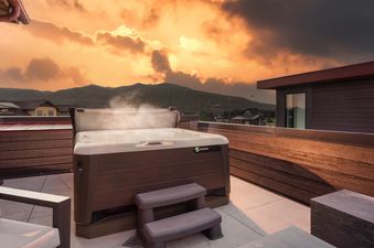 A hot tub with steam rising is on a rooftop deck, overlooking mountains and buildings under a dramatic sunset sky.