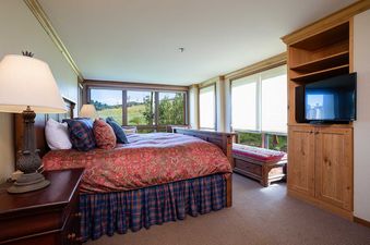 A spacious bedroom at Chateau Chamonix CX121 features a king-size bed with a red and green paisley duvet, a blue plaid bed skirt, and an adjacent nightstand with a lamp. Large windows offer views of the natural landscape, and a wooden entertainment cabine