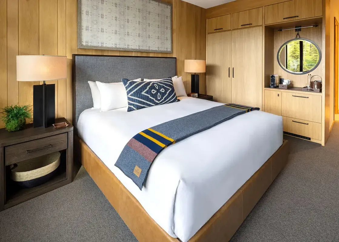 A luxurious king-sized bed with white linens, a blue and white decorative pillow, and a blue and gold throw blanket, flanked by a modern bedside table and a built-in wood wardrobe with a hospitality area.