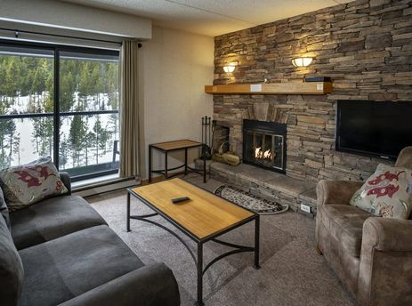 A cozy living room in the Building 4 Two Bedroom Breckenridge Suite with a stone fireplace, comfortable seating, and a large window offering a snowy forest view.