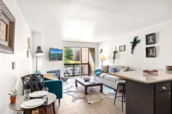 A bright living room in Snowcrest 205 with a leopard print sofa, a teal accent chair, a rustic coffee table, and a dining nook, all bathed in natural light from the balcony doors.