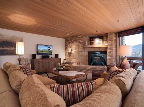 A cozy living room in Chateau Chamonix CX132 features a large sectional sofa, a flat-screen TV, a rustic brick fireplace with a wood-burning stove, and a window with a mountain view.