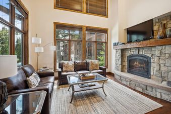 A cozy living room featuring leather sofas, a glass coffee table, and a large stone fireplace with a TV mounted above it, all illuminated by natural light from expansive windows.