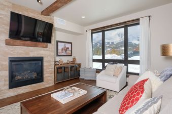 A cozy and modern living room with a fireplace, a large TV, comfortable seating, and a window showcasing a stunning mountain view.