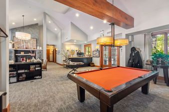 A games room with a pool table, a fireplace, and a reception desk. The pool table is orange, and the fireplace is made of stone. The room is brightly lit with natural light coming from the window and artificial light coming from the recessed ceiling light