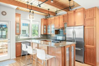 A modern kitchen with light wood cabinetry, granite countertops, stainless steel appliances, and a breakfast bar with white stools.