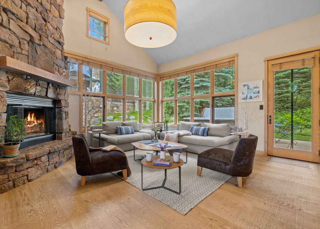 A cozy, sunlit living room at Moose Creek 15 featuring a stone fireplace and forest views through large windows.