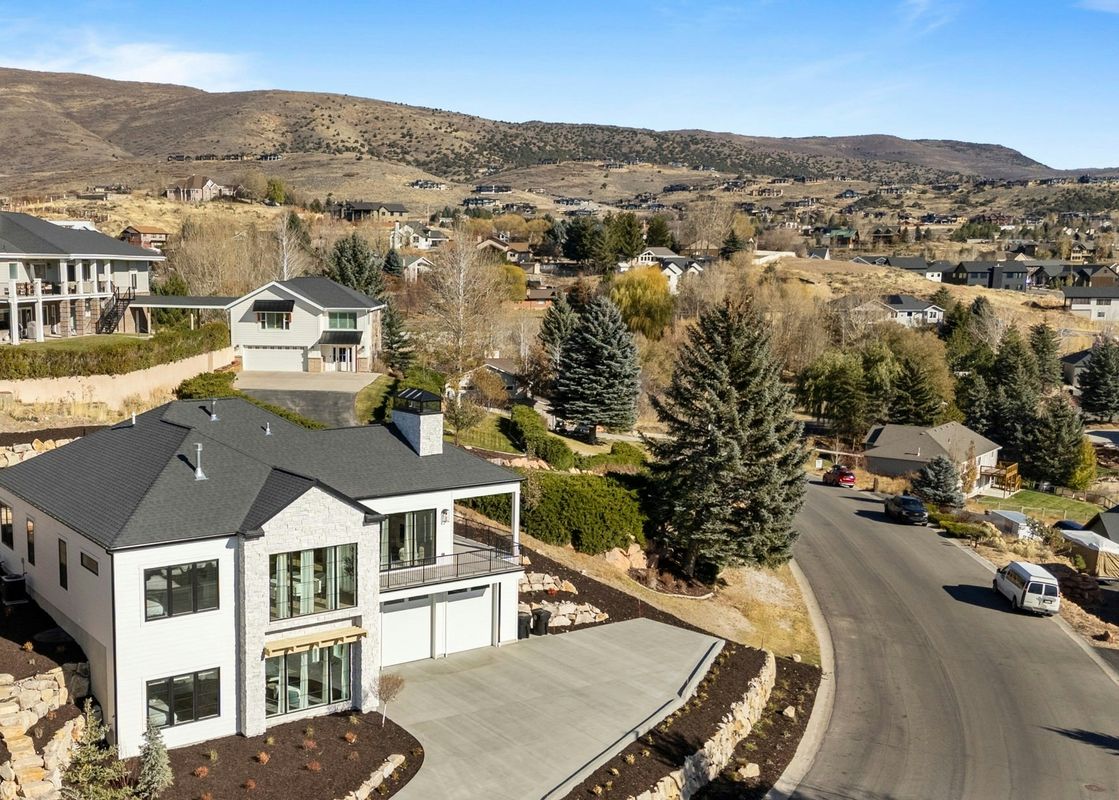 An elevated view of the TimpView Bundle, a modern white and stone house with a dark roof and large windows, situated on a terraced lot with a driveway, double garage, and a balcony. The property is surrounded by landscaped grounds, and evergreen trees