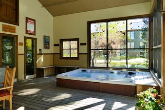 A wooden hot tub sits in a sunlit room with a large window looking out onto a yard and the trees beyond.