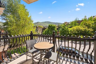 A balcony with a small dining table and two chairs overlooks lush trees and mountains under a blue sky.